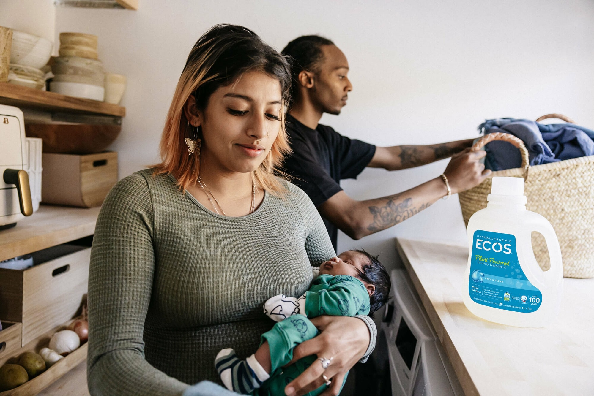 Mom holding newborn baby with dad in background doing laundry and ECOS laundry detergent on counter